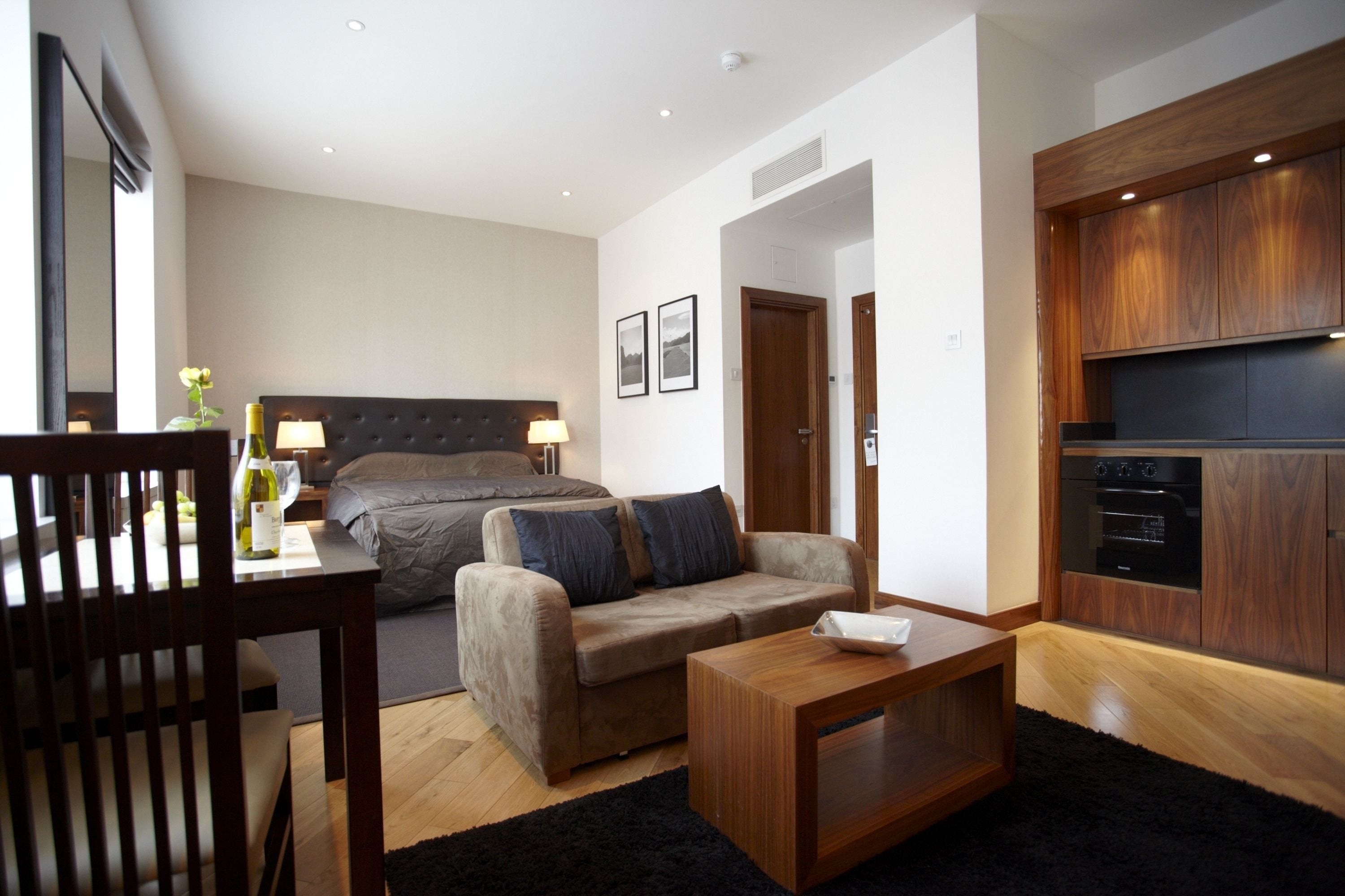 A modern hotel room with a bed, a sofa, a dining table, and a kitchen area.