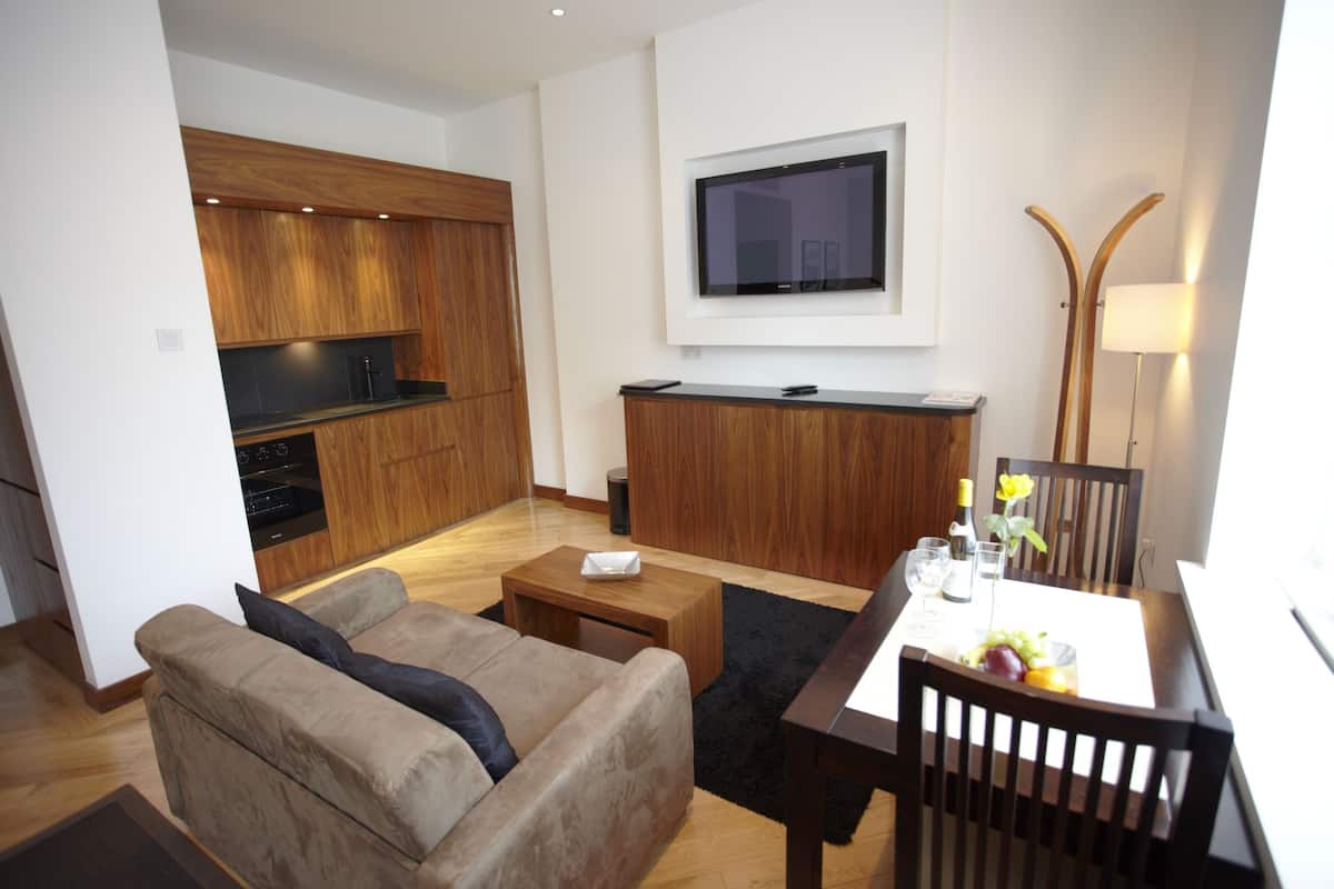A modern living room with a sofa, a dining table with chairs, a TV mounted on the wall, and a kitchen area with wooden cabinets and a stove.