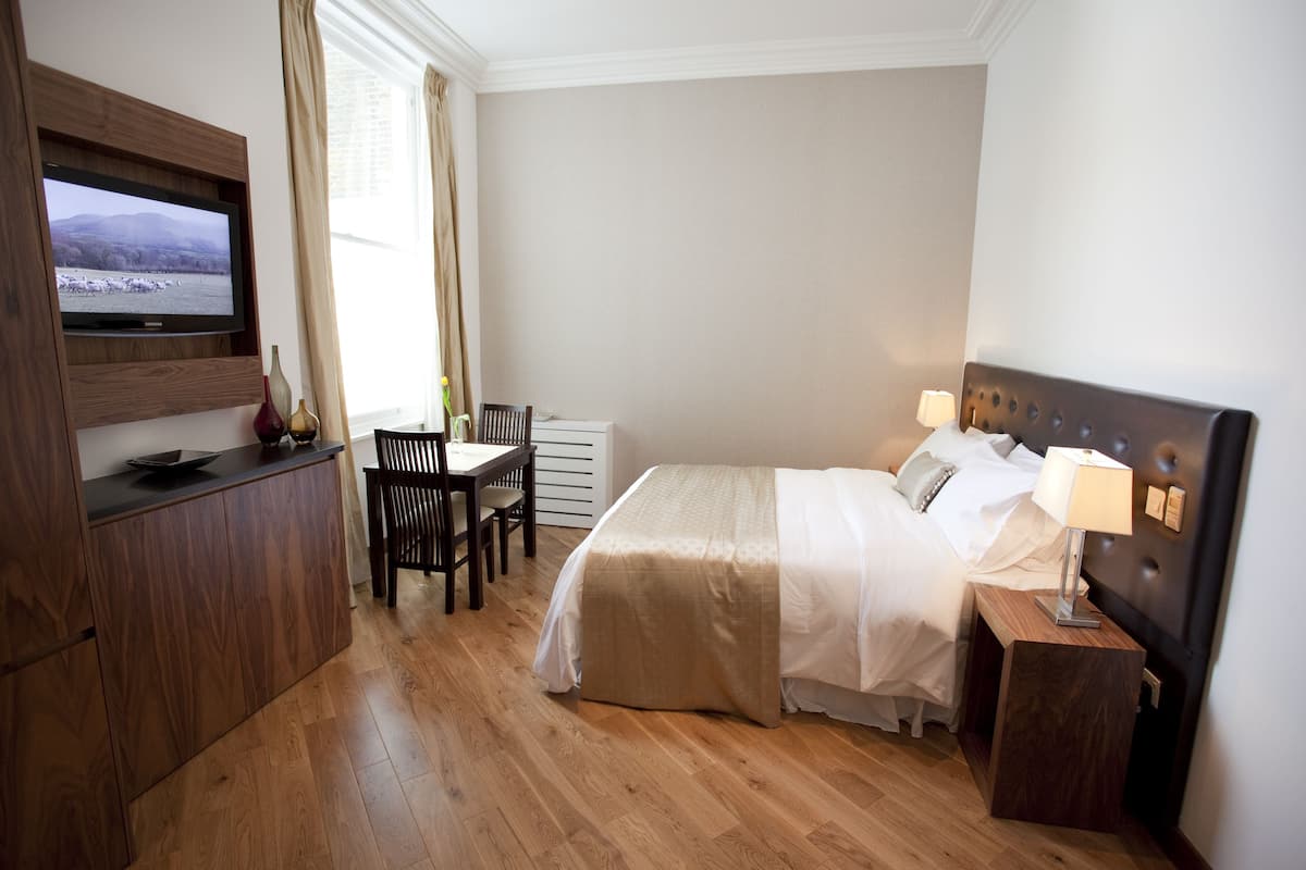 A hotel room with a bed, bedside table, lamp, TV mounted on the wall, wooden floor, and a window with curtains.