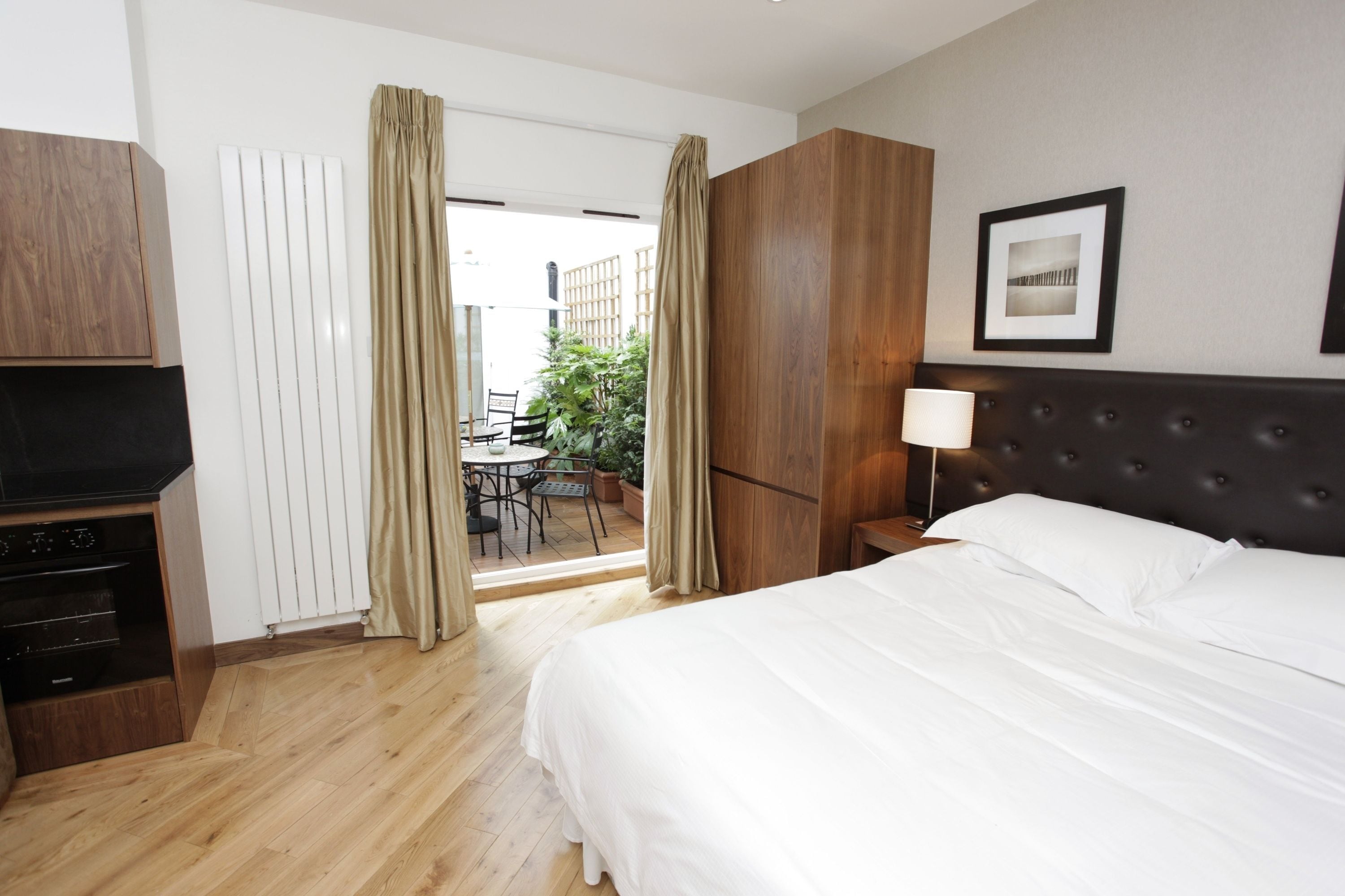 A hotel room with a bed, a TV, a wooden wardrobe, a bedside table with a lamp, and a view of the outside patio.