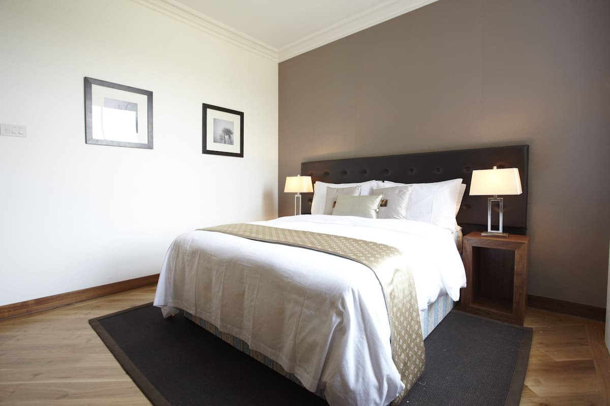 A neatly made bed with a white and beige bedspread, two bedside lamps, and a dark rug on a wooden floor.