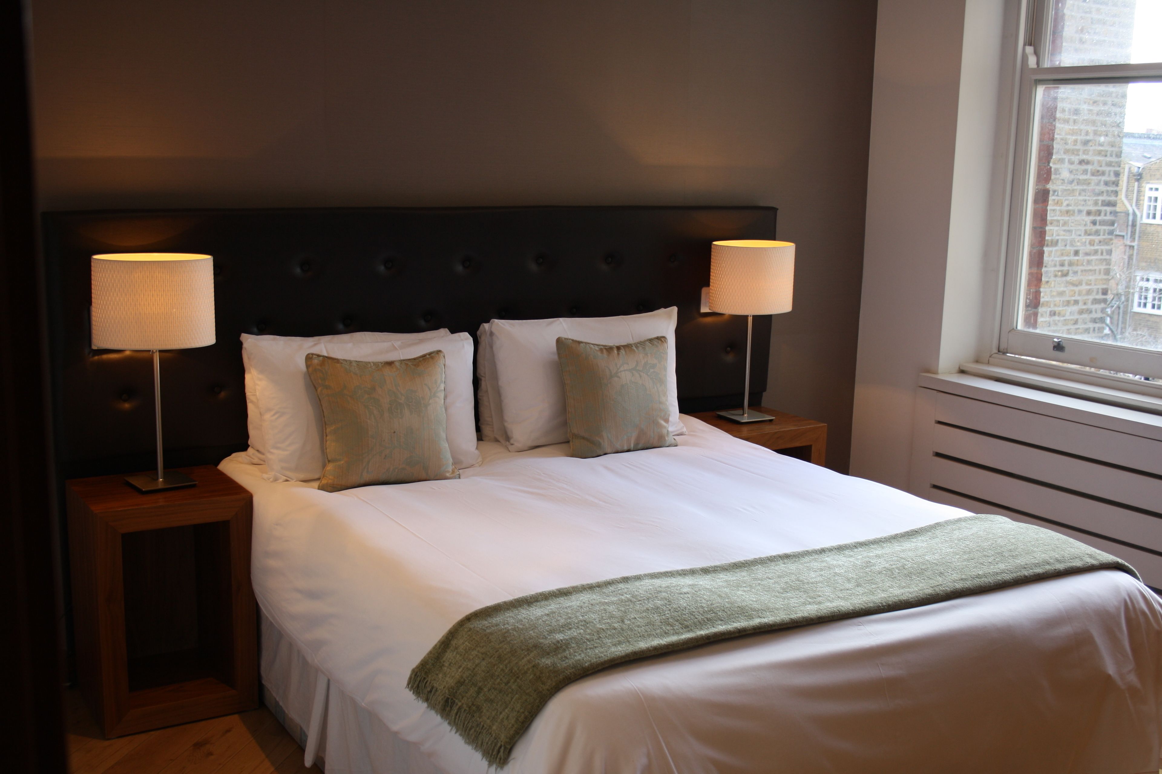A neatly made bed with white linens and a green throw blanket, flanked by two bedside tables with lamps, in a room with a dark headboard and a window with a view of buildings.