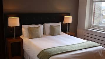 A neatly made bed with white linens and a green throw blanket, flanked by two bedside tables with lamps, in a room with a dark headboard and a window with a view of buildings.