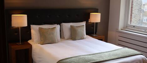 A neatly made bed with white linens and a green throw blanket, flanked by two bedside tables with lamps, in a room with a dark headboard and a window with a view of buildings.