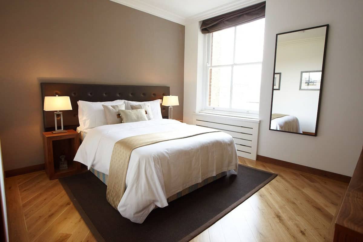 A neatly arranged bedroom with a large bed, two bedside tables with lamps, a window with curtains, and a wall-mounted mirror.