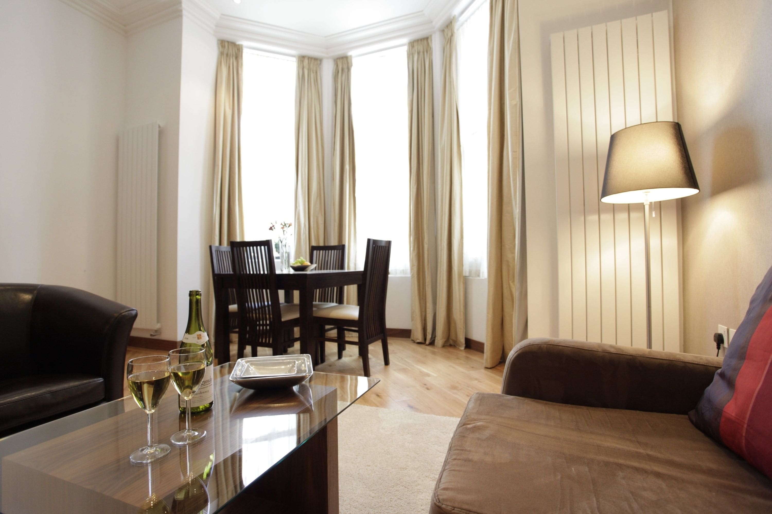 A modern living room with a glass-top table, two wine glasses, a bottle, and a lamp.