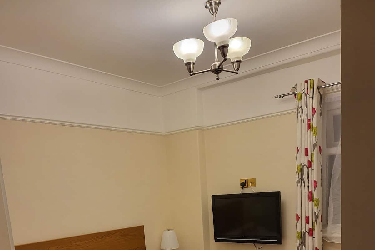 Quarto de hotel com uma cama grande, televisão de tela plana, cabeceira de madeira, mesa de cabeceira com luminária e janela com cortinas florais.