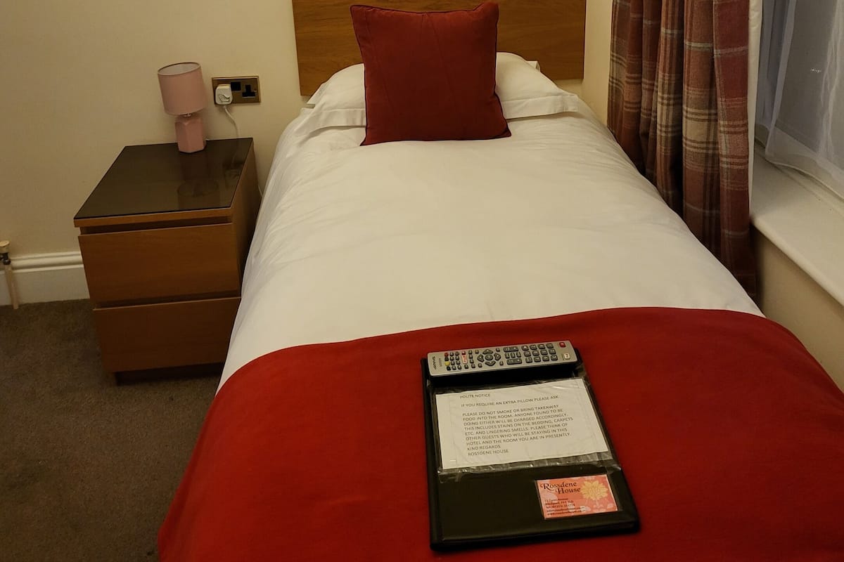 Quarto de hotel com cabeceira de madeira, travesseiro vermelho, colcha branca, manta vermelha, controle remoto e mesa de cabeceira com abajur.