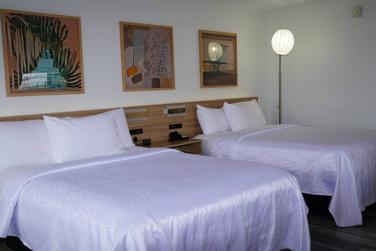 A hotel room with two beds, a nightstand, a lamp, and artwork on the wall.