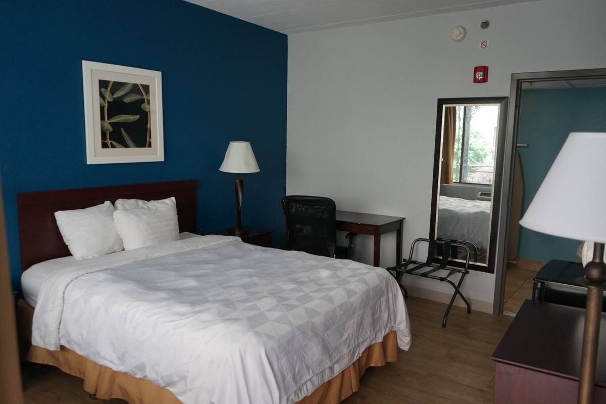 A hotel room with a bed, a desk, a chair, a lamp, and a mirror.