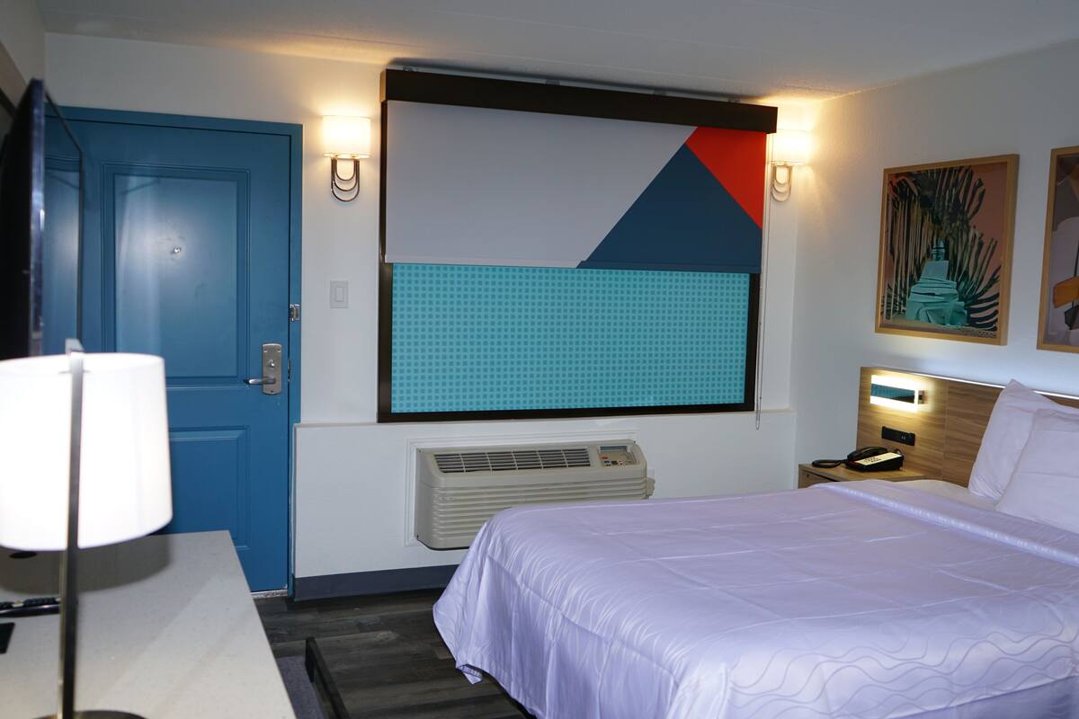 A hotel room with a bed, a desk with a lamp, a phone, a wall-mounted heater, and a door.
