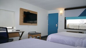 Habitación de hotel con dos camas, un escritorio con una silla, un televisor montado en la pared, una puerta azul y una ventana con vistas a una cerca.