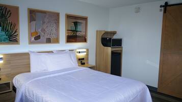A neatly made bed with white linens, a wooden headboard, and a nightstand with a lamp. There are framed pictures on the wall and a sliding door to the right.