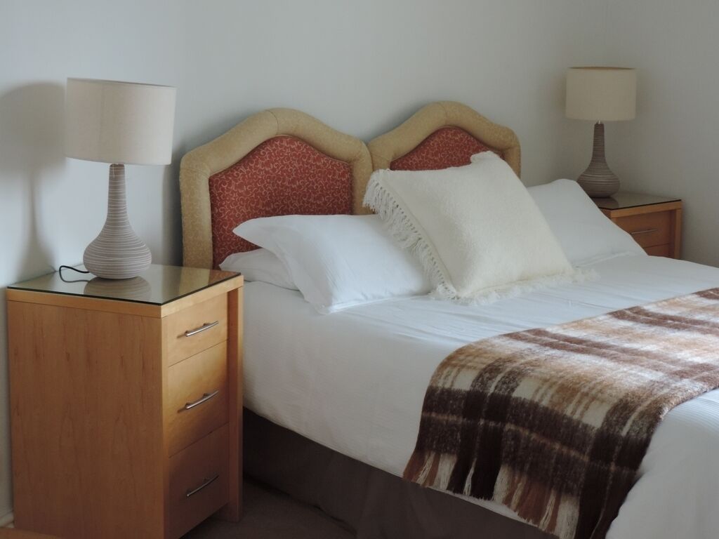 A neatly made bed with white linens and a plaid throw blanket, flanked by two bedside tables with lamps, each with a wooden base and a lamp with a beige lampshade.
