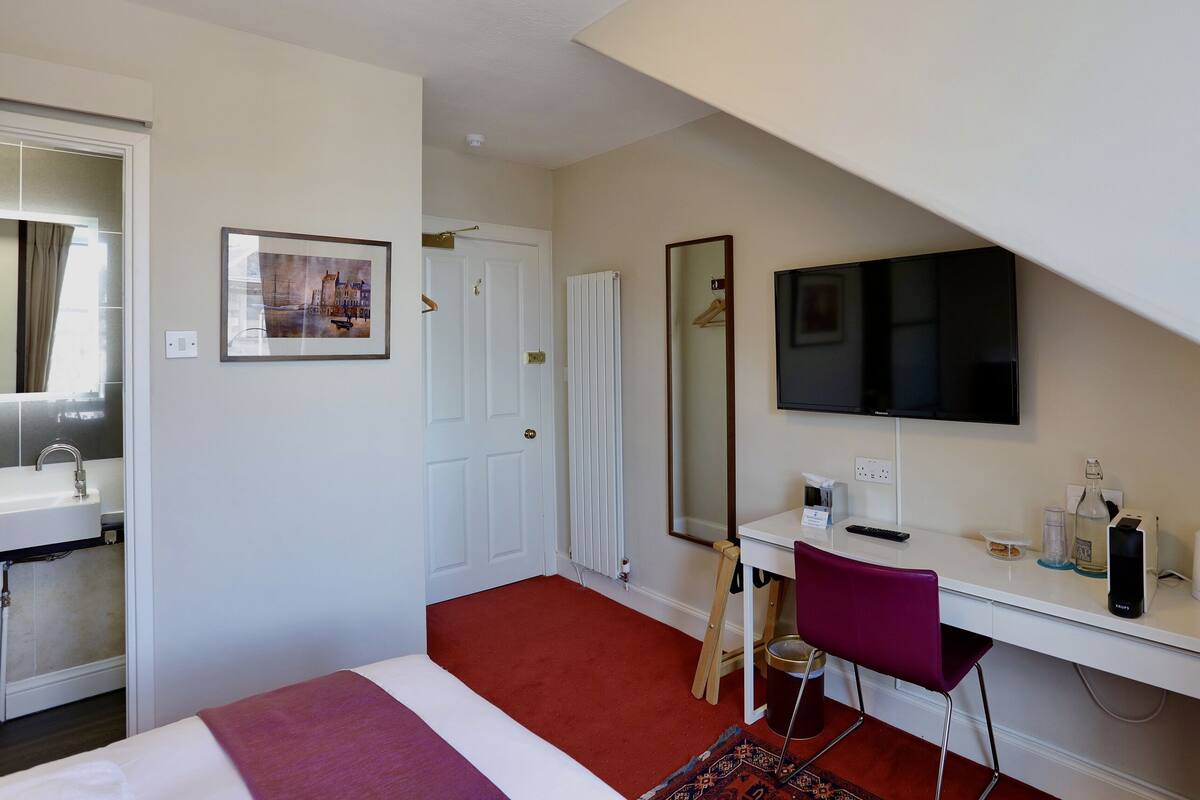 A hotel room with a bed, a desk with a computer, a TV, a mirror, a window, and a door.