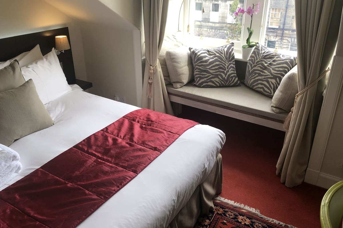 A hotel room with a bed, a window seat with cushions, and a view of buildings outside.