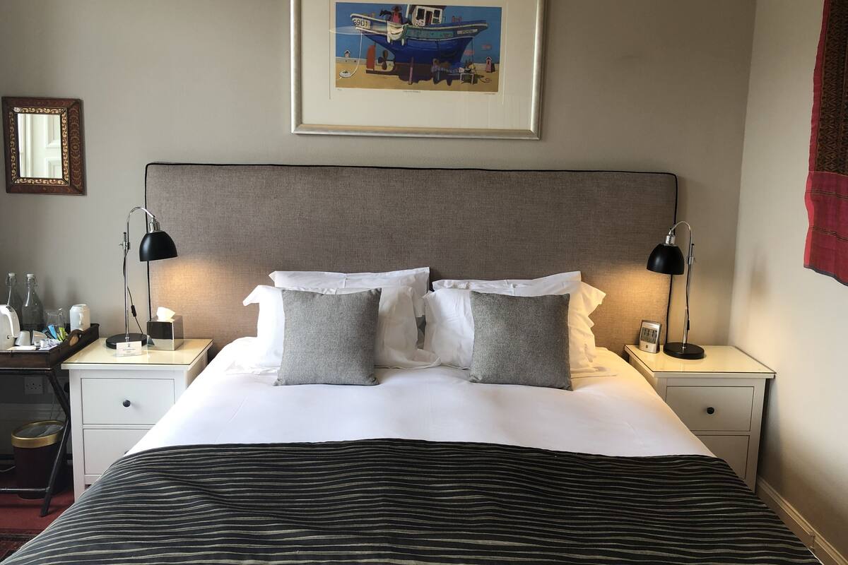 A neatly made bed with white and grey pillows, a striped blanket, and a framed painting of a boat above it. There are two bedside tables with lamps and a small clock.