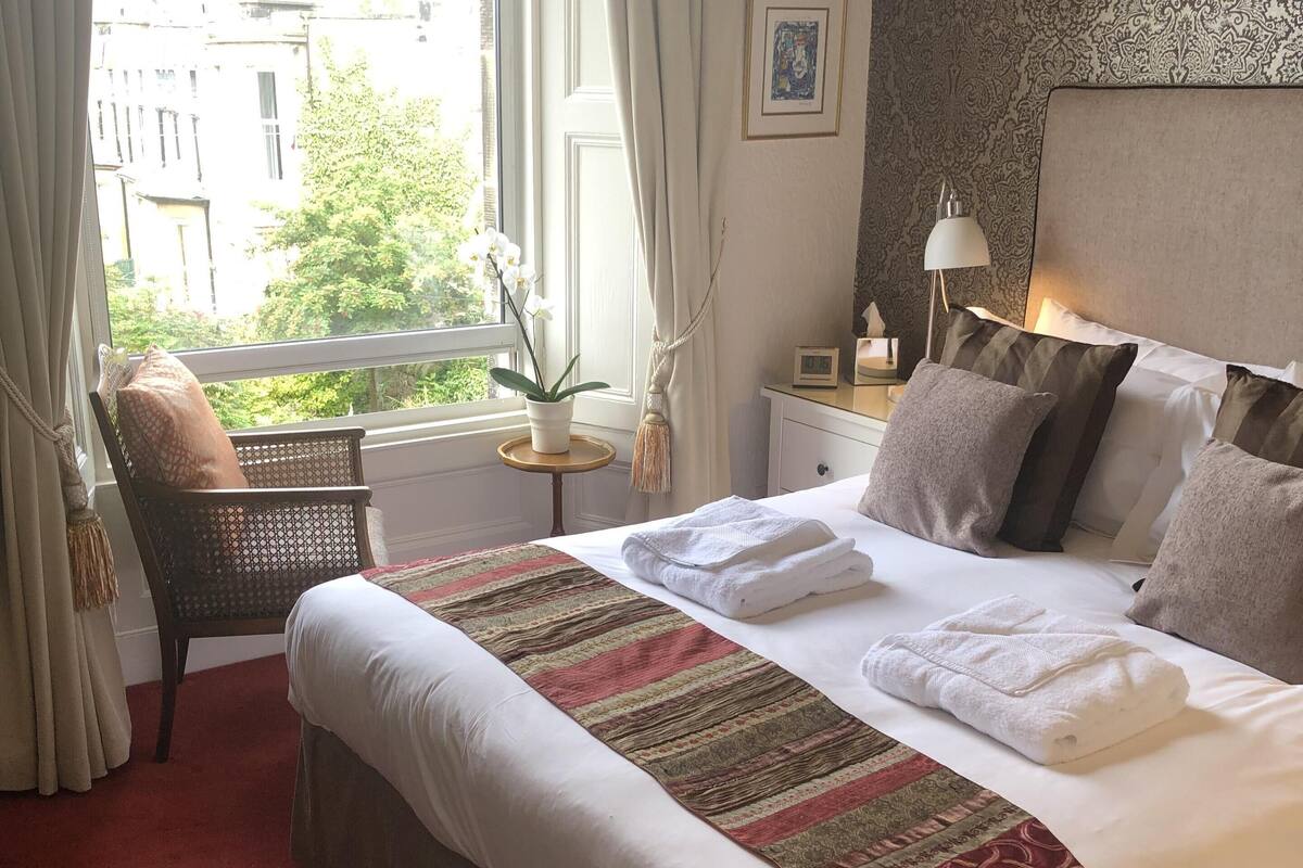 A neatly made bed with a striped blanket, a red patterned rug, a chair with a cushion, a small table with a plant, and a window with curtains.