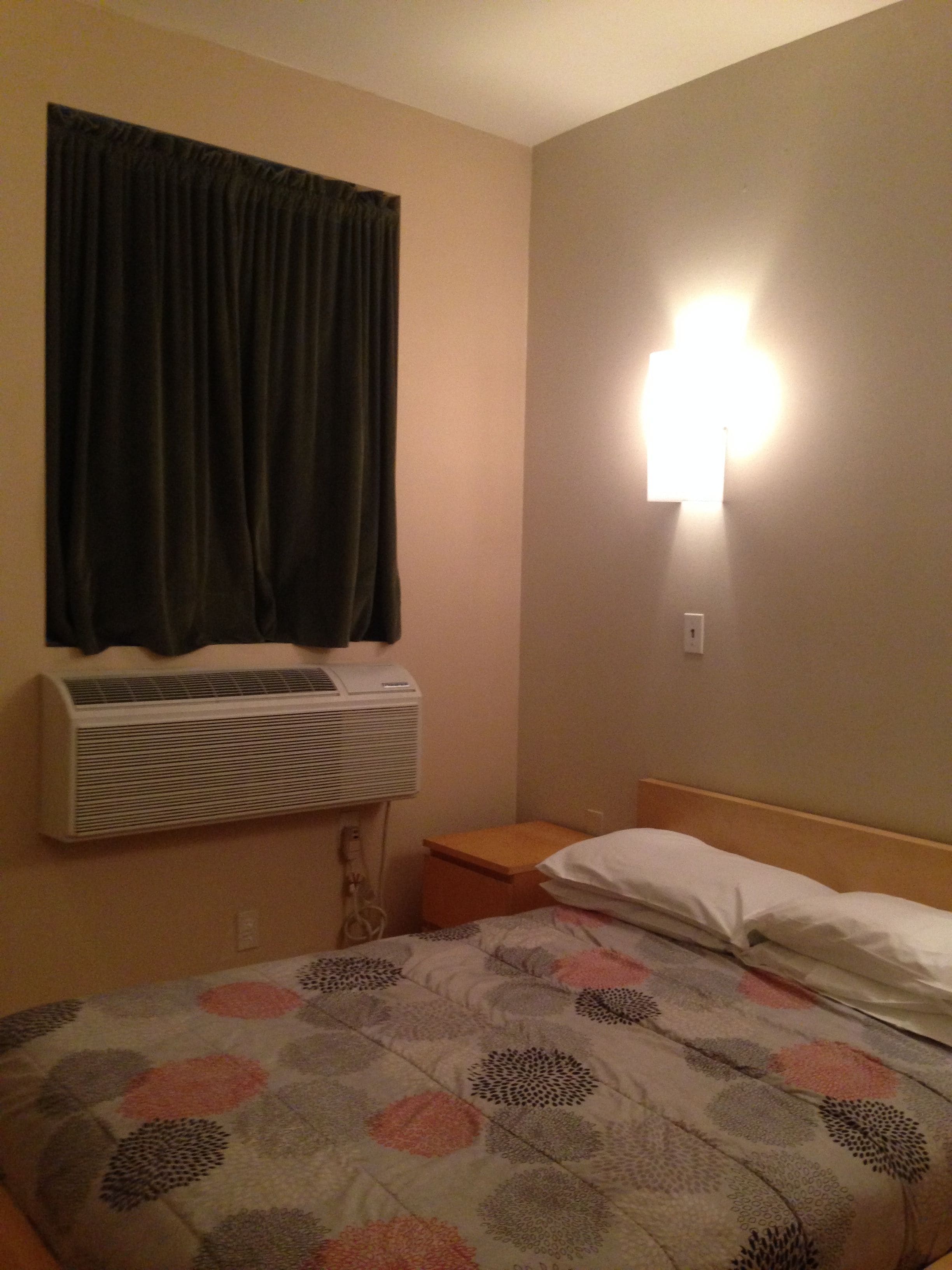 Una habitación de hotel con cama, aire acondicionado de pared, mesita de noche y luz de pared.