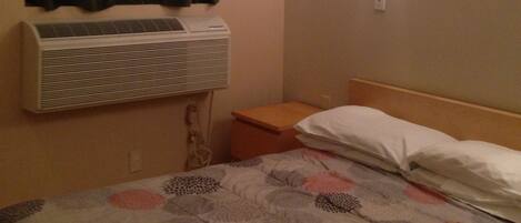 Una habitación de hotel con cama, aire acondicionado de pared, mesita de noche y luz de pared.