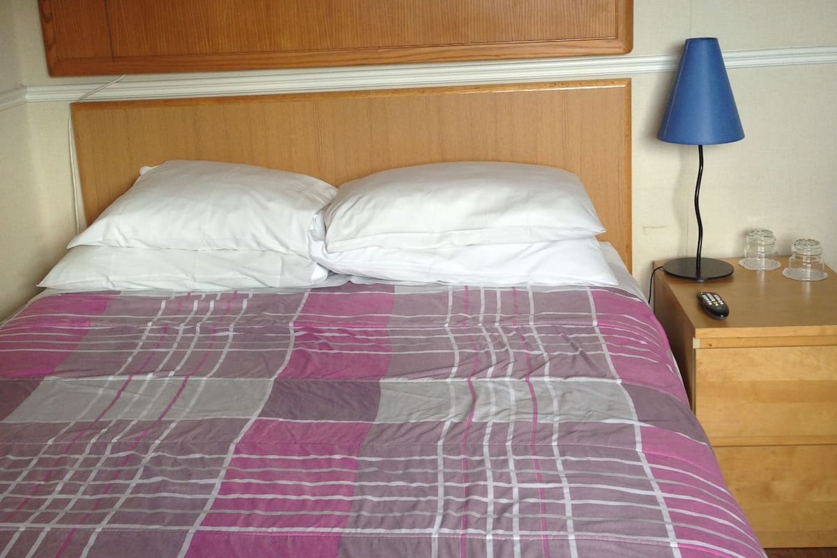 A hotel room with a bed, a wooden headboard, a nightstand with a lamp, and a remote control.