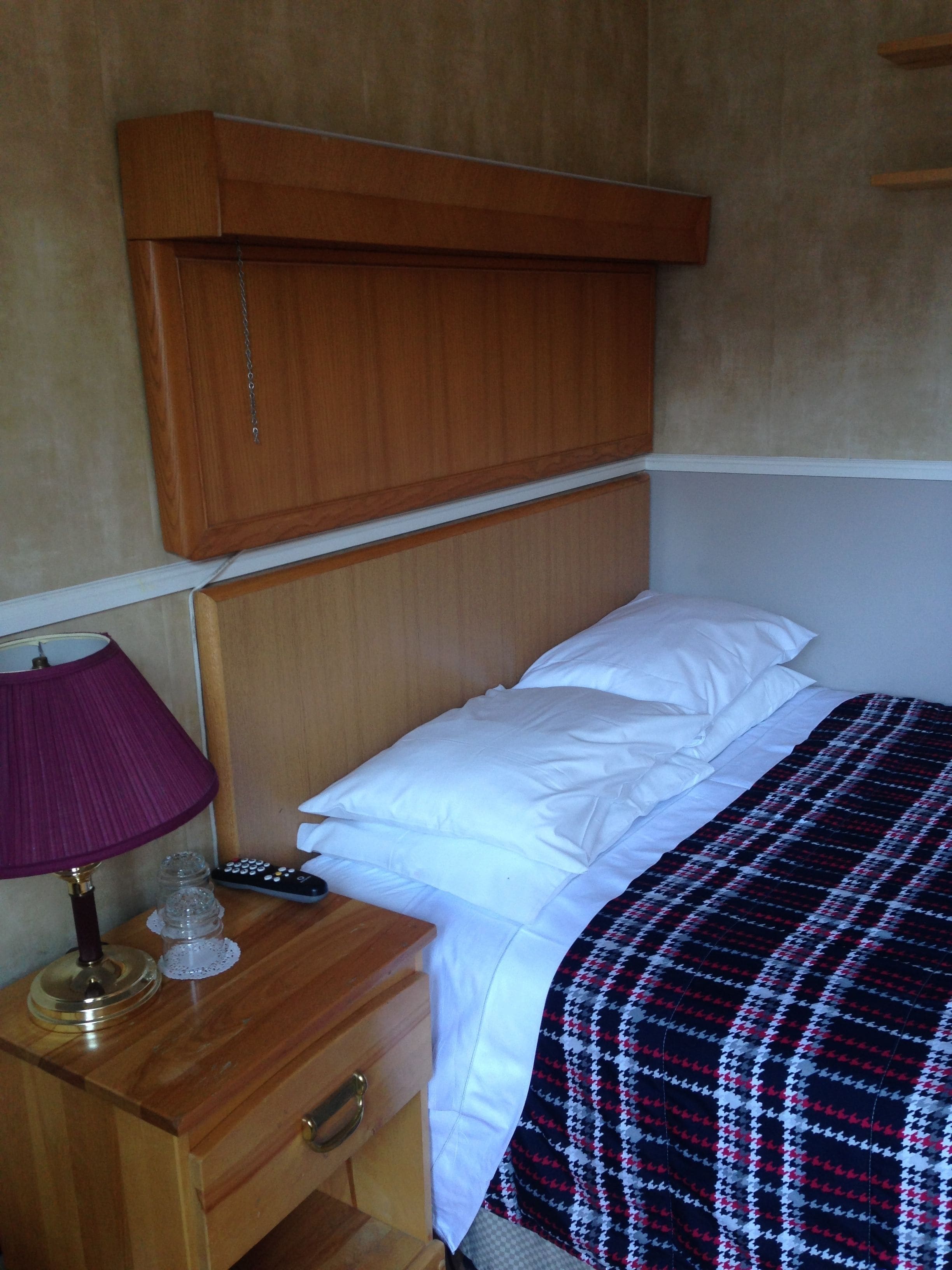Un dormitorio con cabecera de madera, cama con sábanas blancas, manta escocesa, mesita de noche con lámpara, control remoto y una botella decorativa.