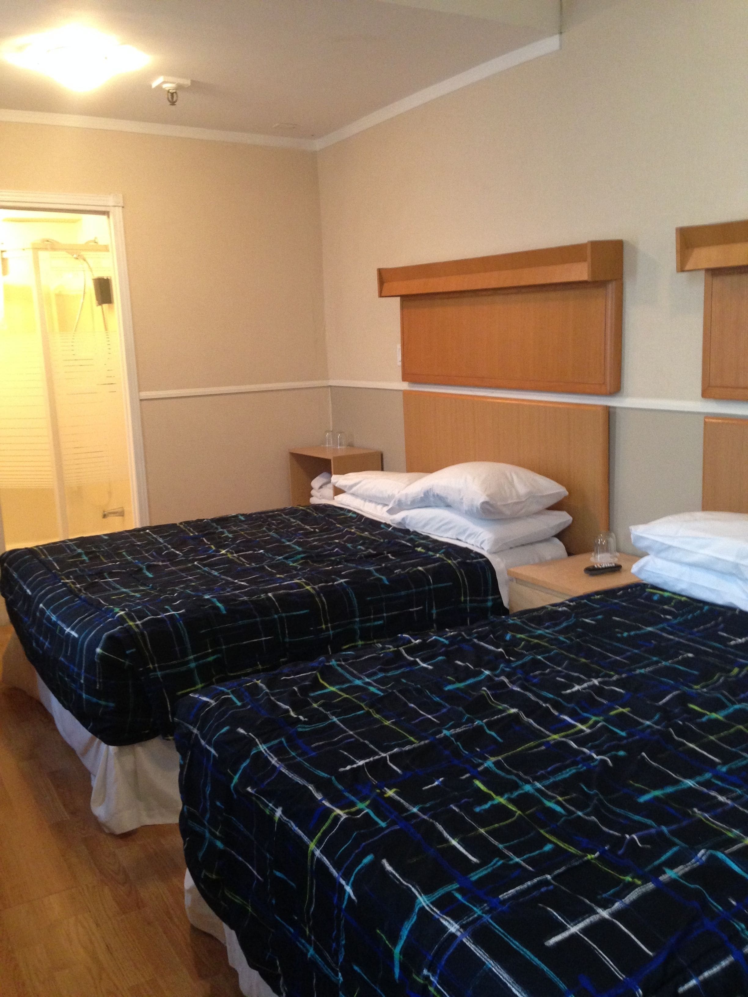 A hotel room with two beds, wooden headboards, a bedside table with a lamp, and a door with a window.