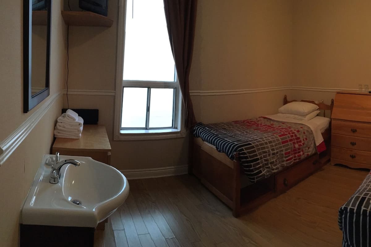 A small, well-lit bedroom with two single beds, a wooden dresser, a bedside table, and a bathroom with a sink and mirror.