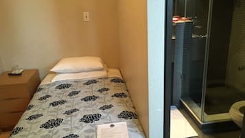 A small, single-bed room with a patterned bedspread, a bedside table, and a shower area visible through a glass door.