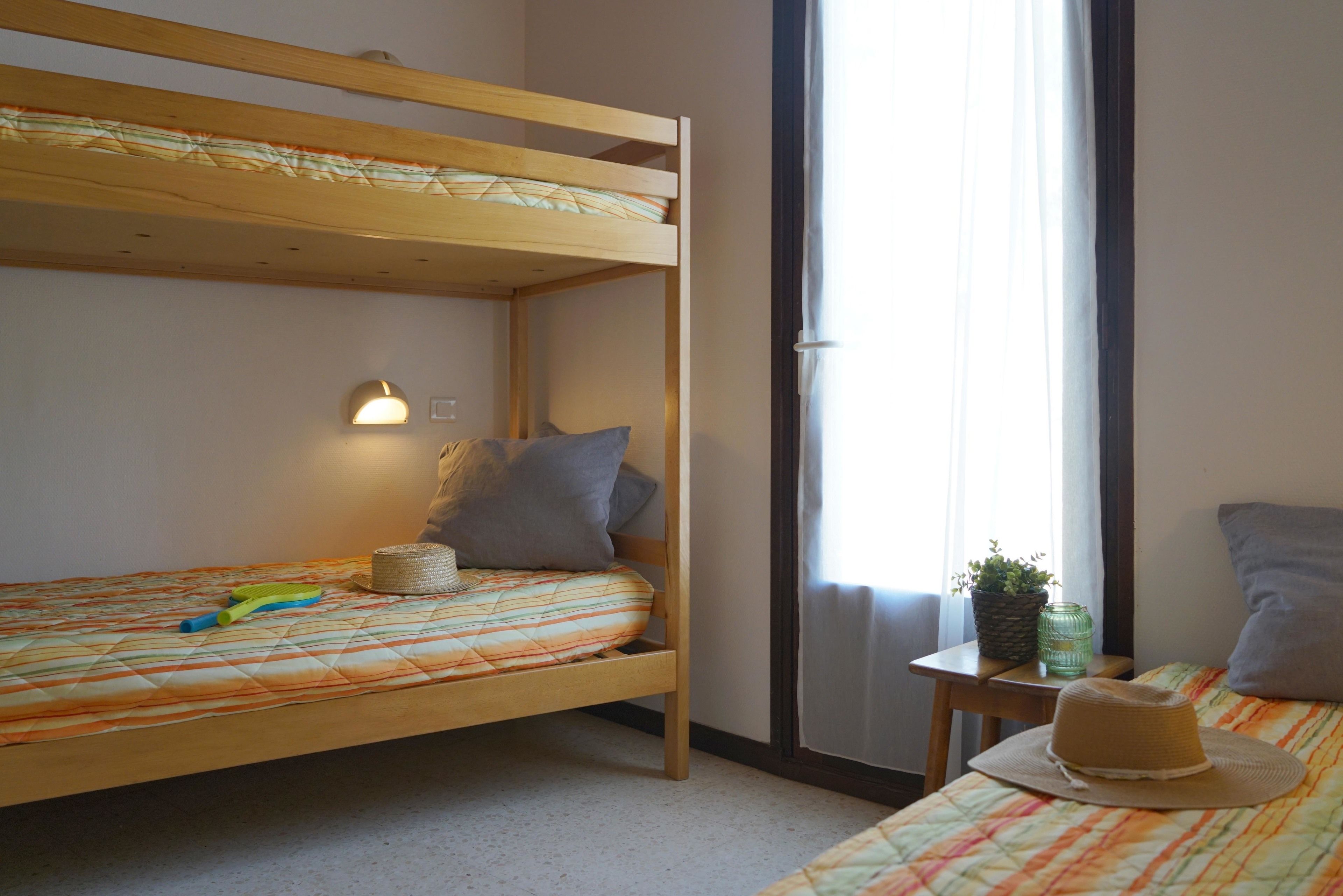 A bunk bed with a striped bedspread, a small bedside table with a plant and a candle, and a window with curtains.