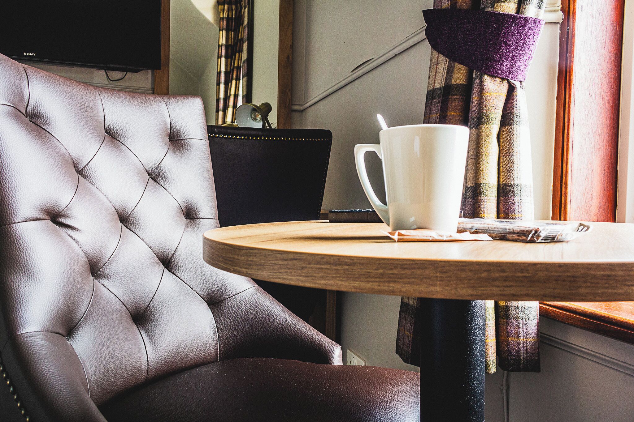 A tufted leather chair, a white mug, and a plaid curtain in a room with a television and a window.