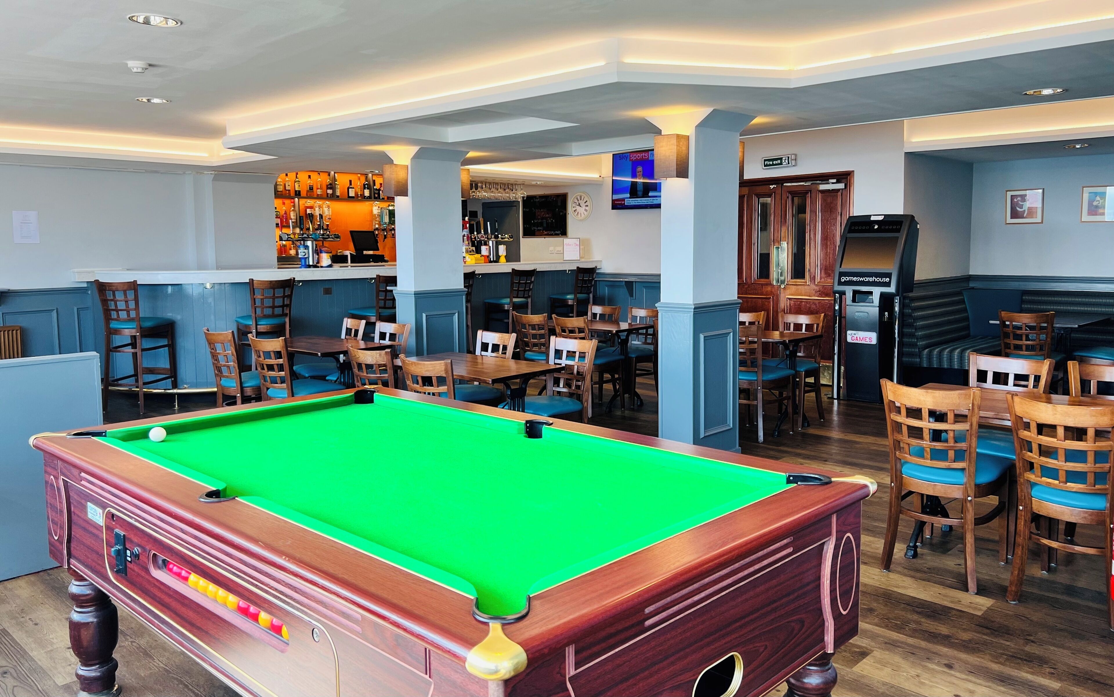 A pool table in a room with multiple seating areas, a bar with a variety of bottles, and a television displaying a news channel.
