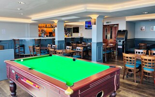 Mesa de bilhar em um espaço com vários cantos de estar, um bar com diversas garrafas e uma televisão exibindo um noticiário.