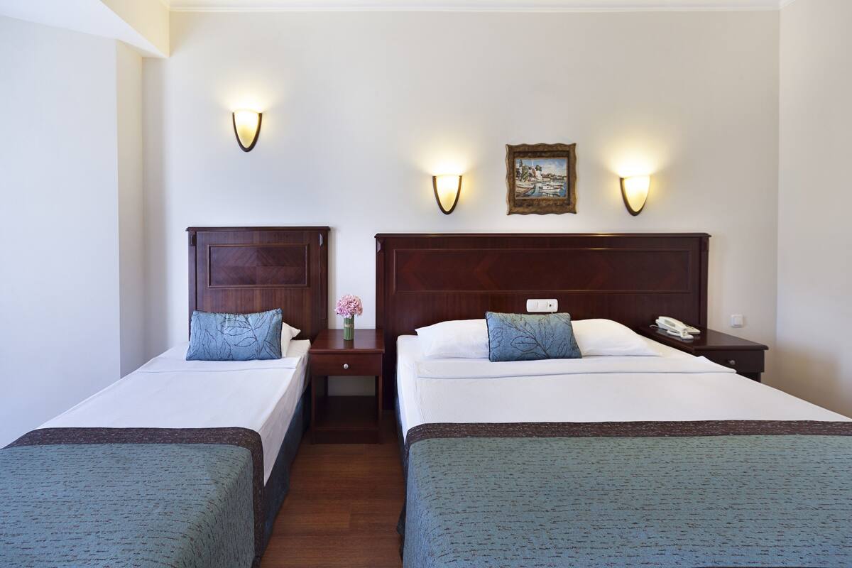 A hotel room with two single beds, each with a wooden headboard, white linens, and a blue throw blanket. There are wall-mounted light fixtures and a small table with a vase of flowers between the beds.