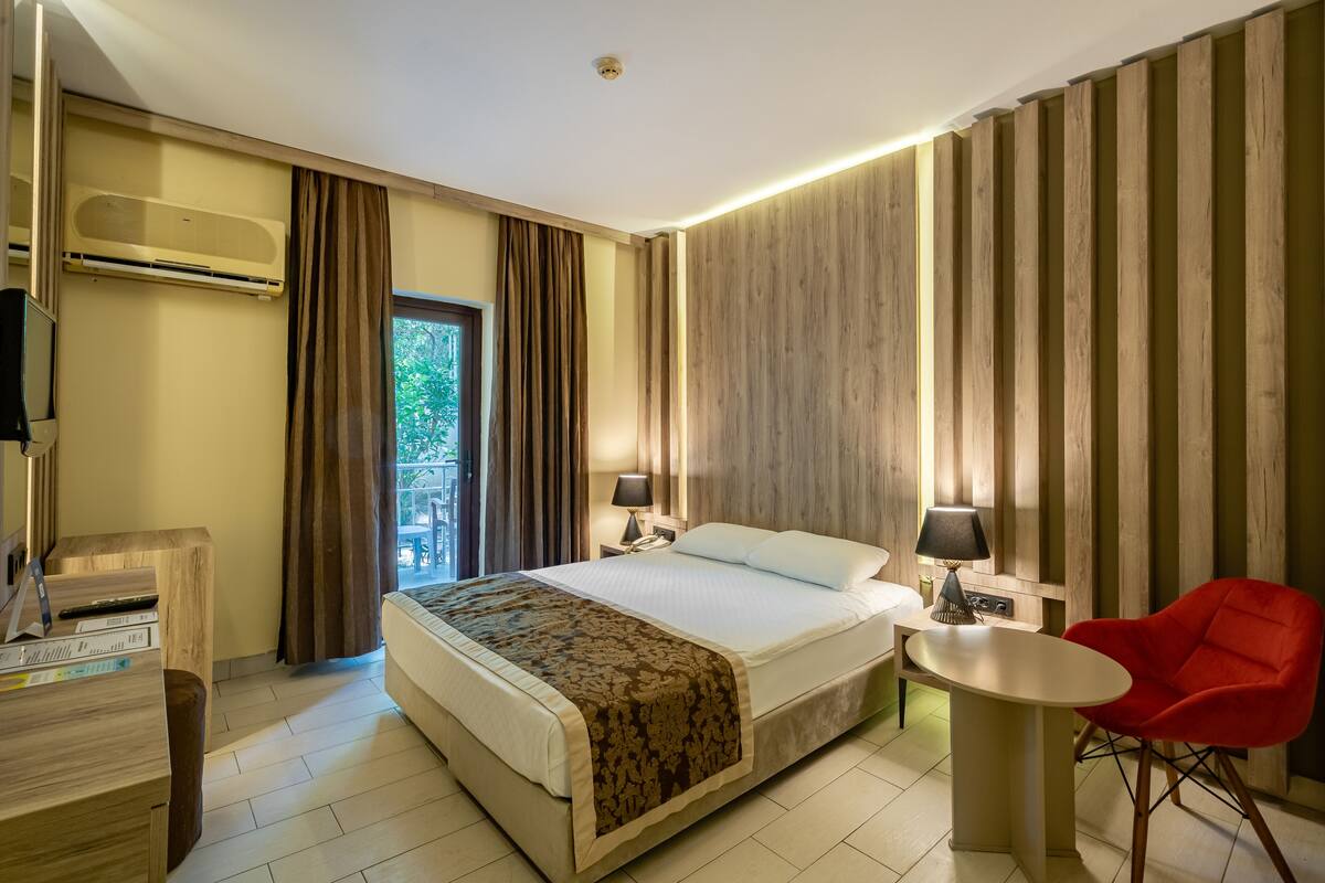 A hotel room with a large bed, bedside tables with lamps, a red chair, and a view of greenery outside.