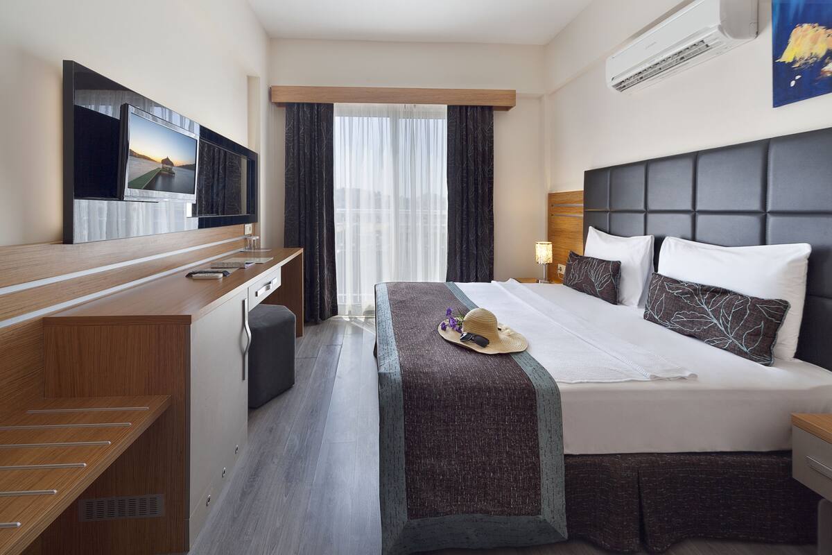A hotel room with a bed, a desk with a flat-screen TV, a window with curtains, and an air conditioning unit.