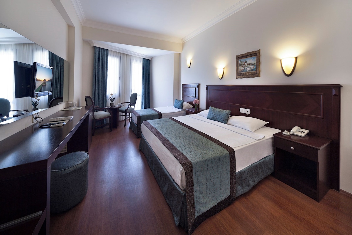 Quarto de hotel com uma cama grande, uma escrivaninha com computador, uma cadeira, uma mesinha e uma televisão.
