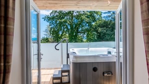 A modern outdoor hot tub area with a wooden deck, a metal chair, and a glass door leading to a lush green landscape. - Orestone Manor (Torquay)