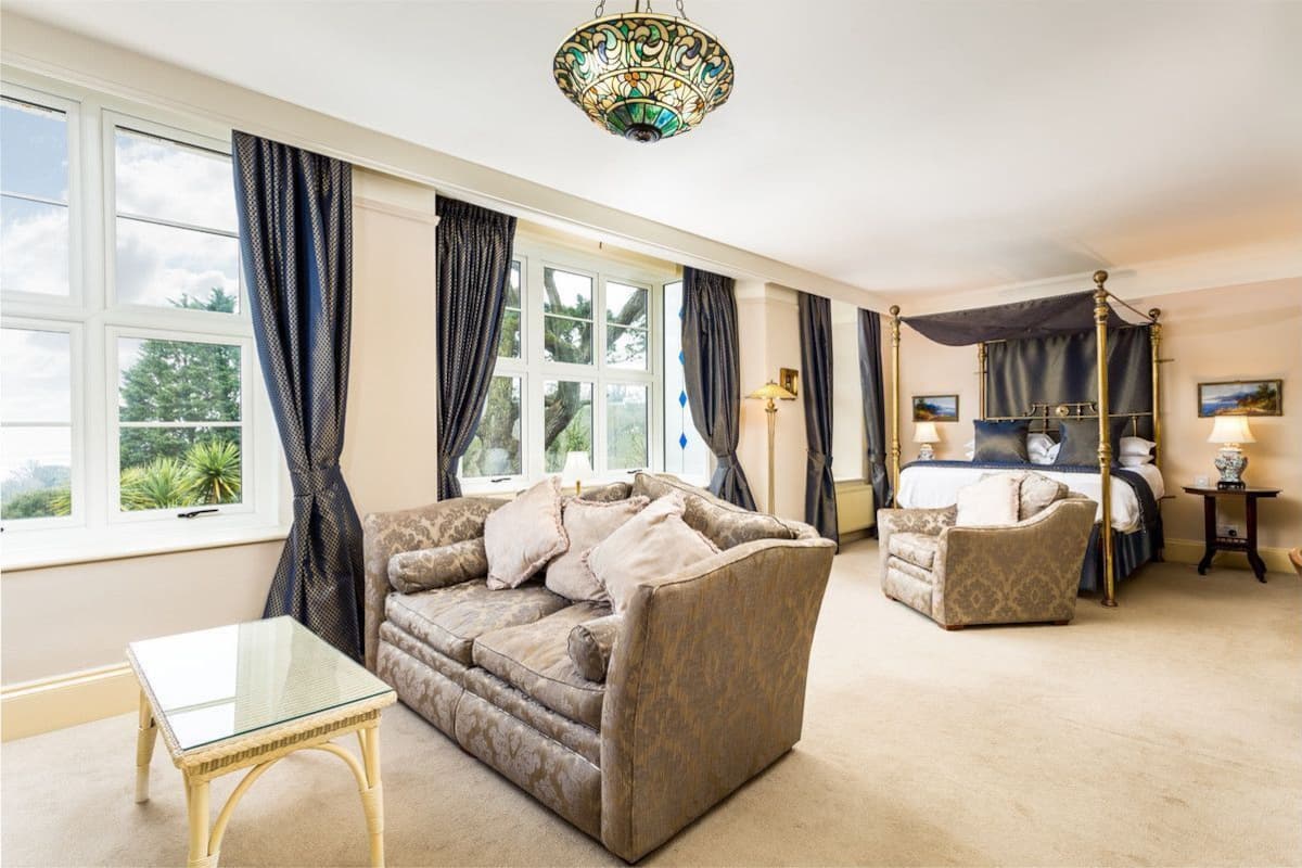 Ein Zimmer mit großem Fenster, einem Himmelbett, einer Couch, einem Sessel und einem Glastisch.