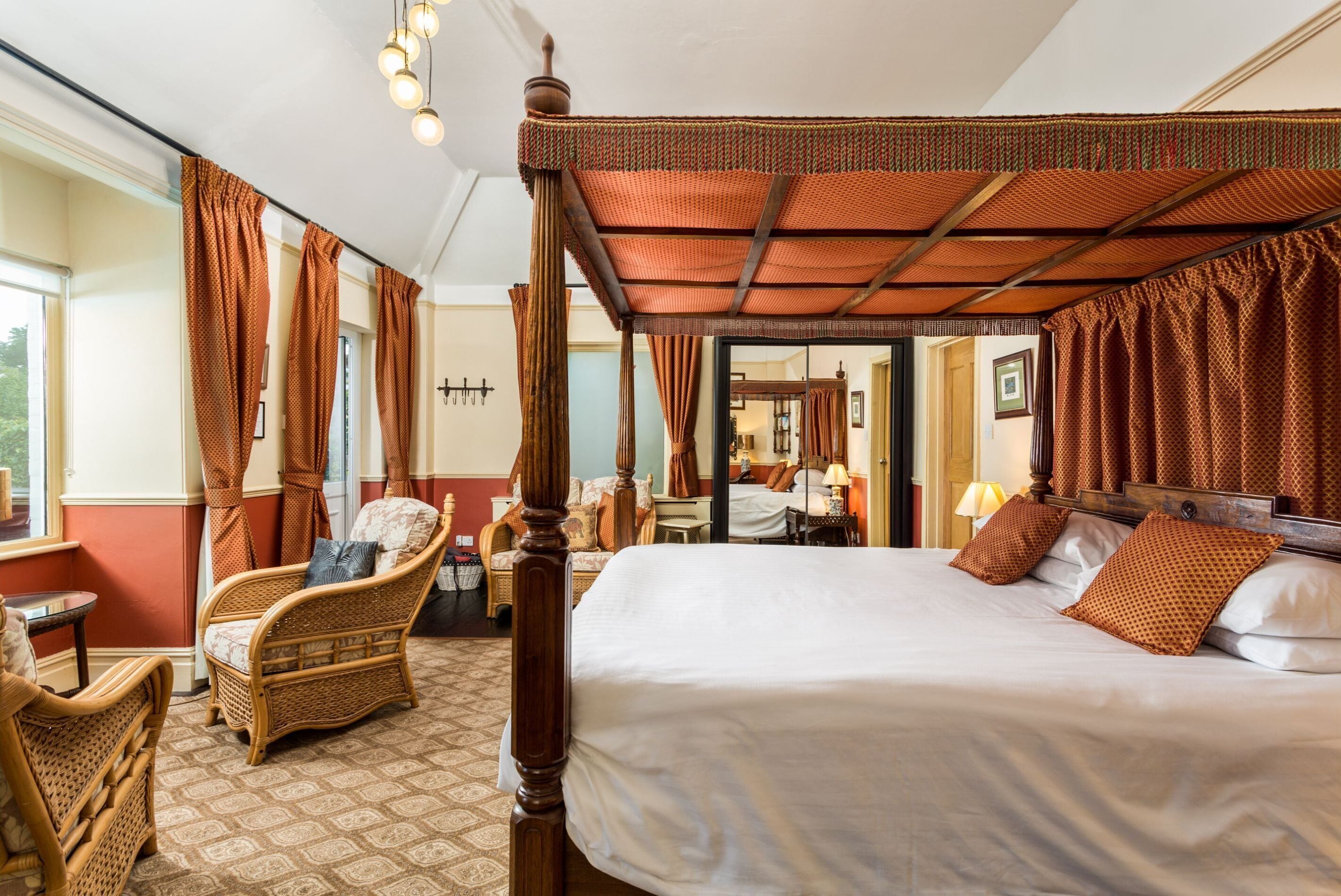 Una cama con dosel y cuatro columnas, un sillón de mimbre y una mesita en una habitación con alfombra estampada.