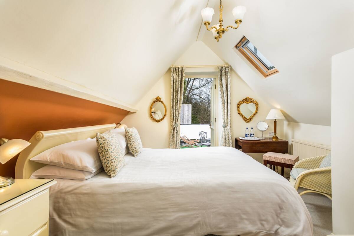 Ein Schlafzimmer mit einem großen Bett, einem Nachttisch, einer Kommode, einem Sessel, einem Schreibtisch mit Lampe und einem Fenster mit Vorhängen.