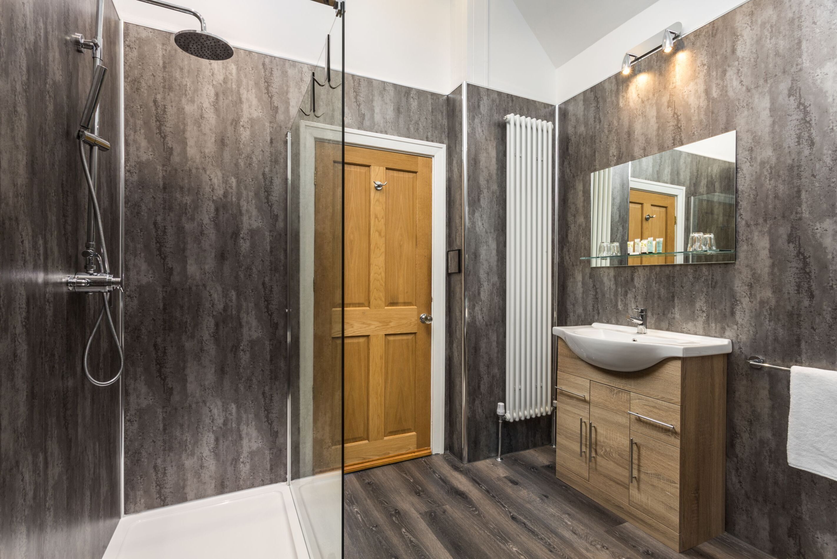 Un baño moderno con ducha, puerta de madera, lavamanos con superficie de vidrio y un espejo grande.
