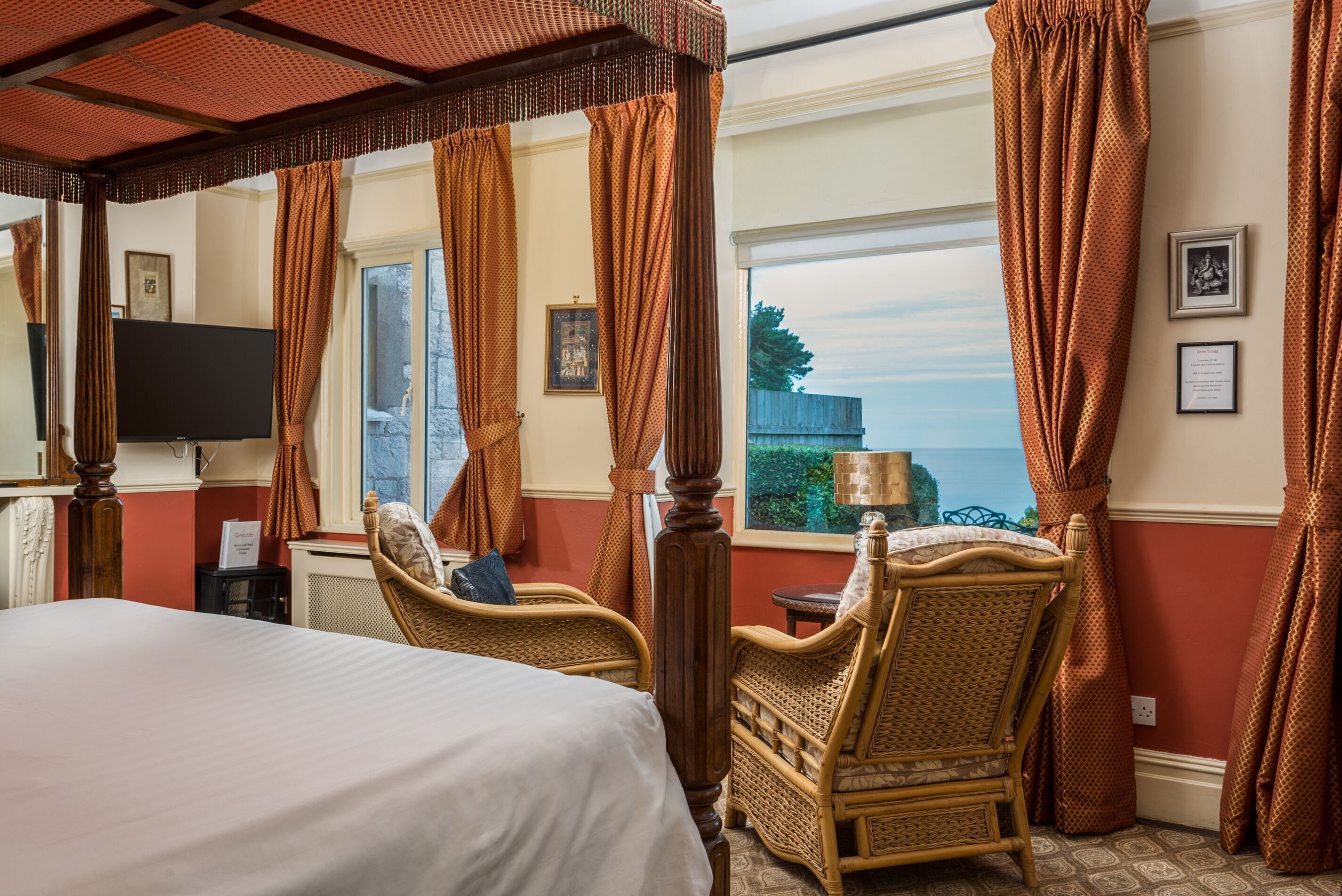 Habitación de hotel con cama con dosel, sillas de mimbre, televisión y vistas al exterior.