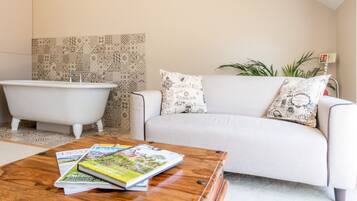 Une salle de bain moderne avec une baignoire sur pieds, un canapé blanc agrémenté de coussins à motifs et une table basse en bois avec des magazines.