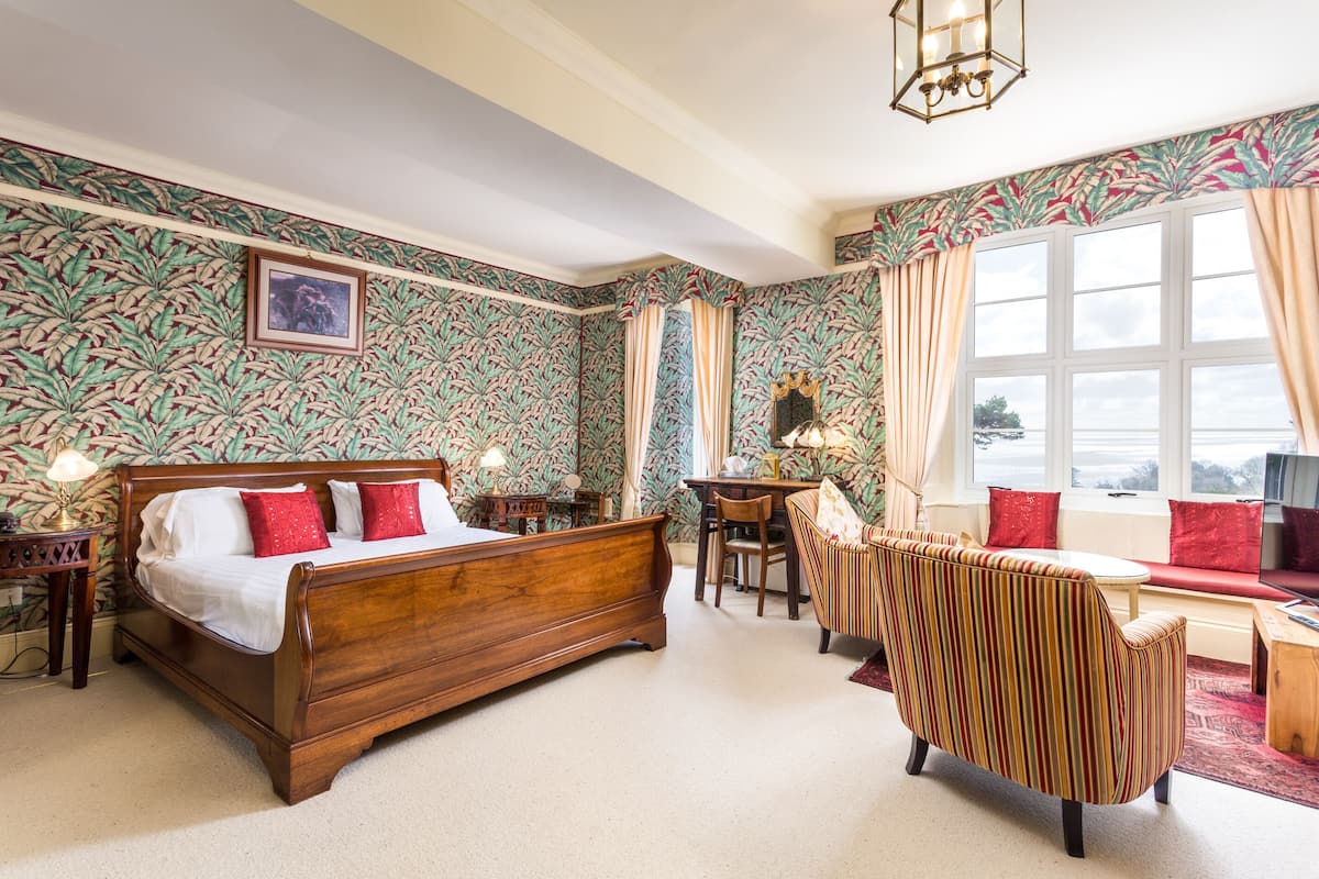 Ein Schlafzimmer mit einem hölzernen Bett, einem gestreiften Sessel, einem Schreibtisch und einem Fenster mit Vorhängen.