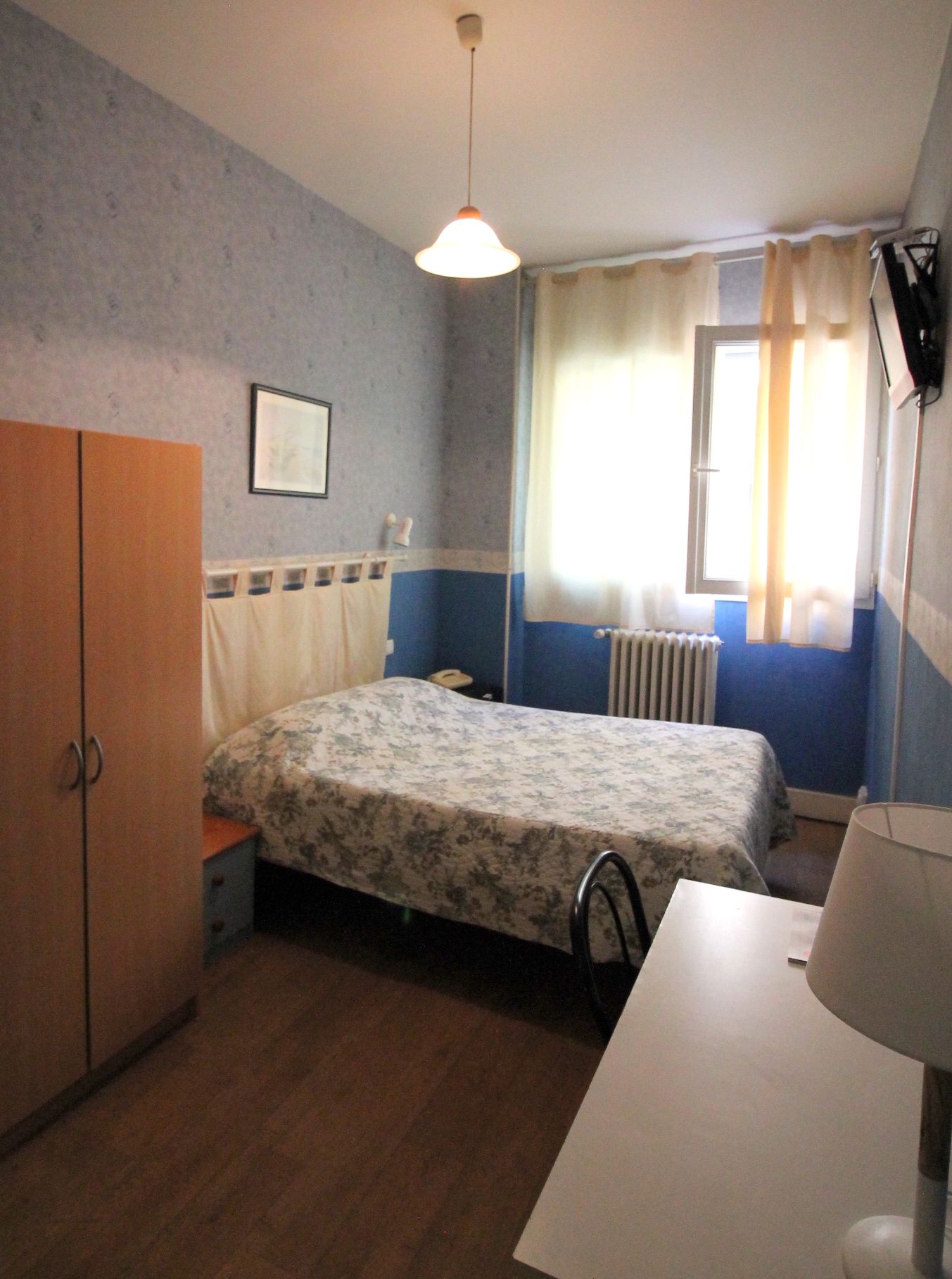 A bedroom with a bed, a wardrobe, a desk, a radiator, and a window with curtains.