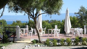 Un patio al aire libre con mesas y sillas blancas, olivos y vistas al mar.