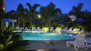 Una piscina con sillas de descanso y sombrillas, rodeada de palmeras.
