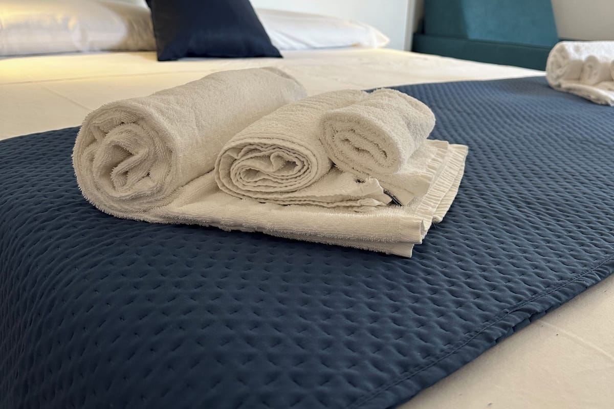 A neatly made bed with a rolled white towel on a blue quilted bedspread, a dark blue pillow, and a white headboard in a hotel room.