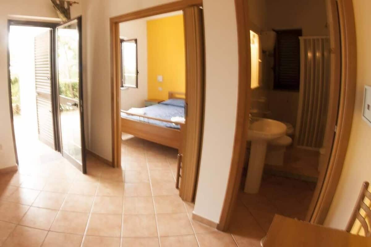 A corridor leading to a bedroom with a bed, a bathroom with a sink and toilet, and a view of the outside.